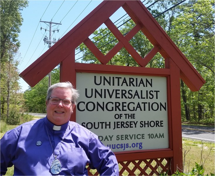 photo of Rev Dawn Fortune, smiling in purple clerical garb, standing in the sunshine in front of the congregations signage